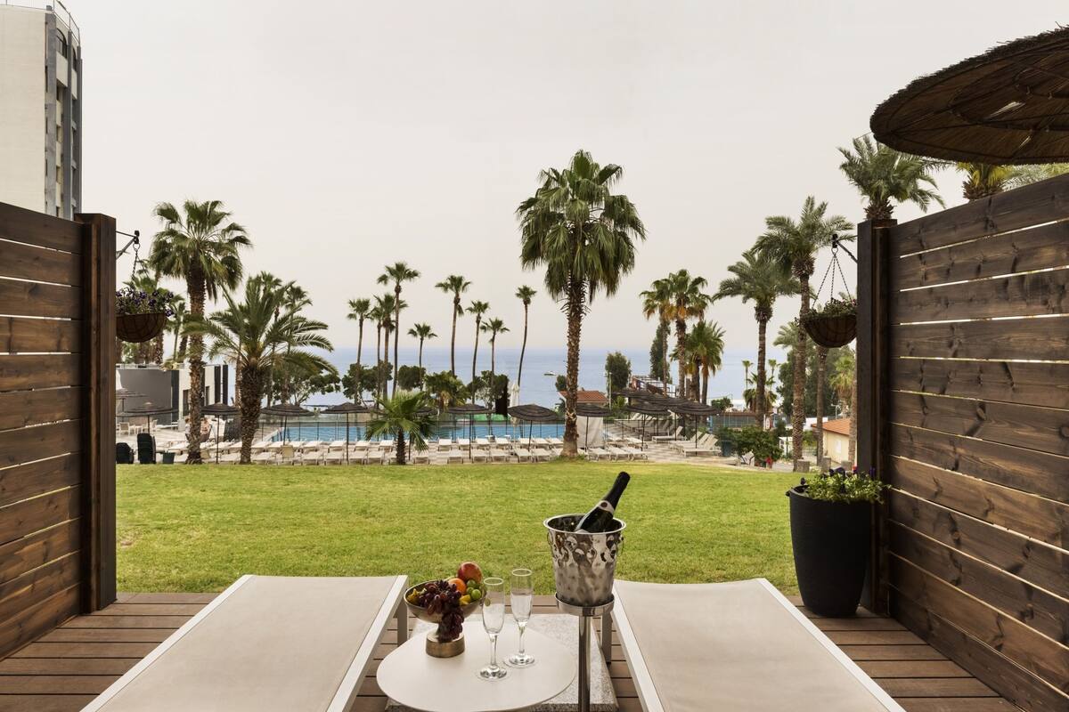 A patio with a table set for two, a fruit bowl, and a bottle of champagne. The view includes a pool and palm trees.