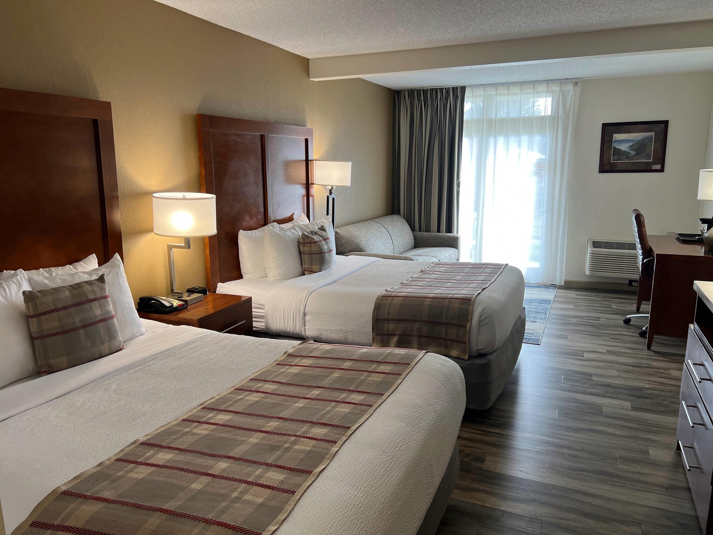 Habitación de hotel con dos camas, un escritorio, una silla, una lámpara y un cuadro en la pared.