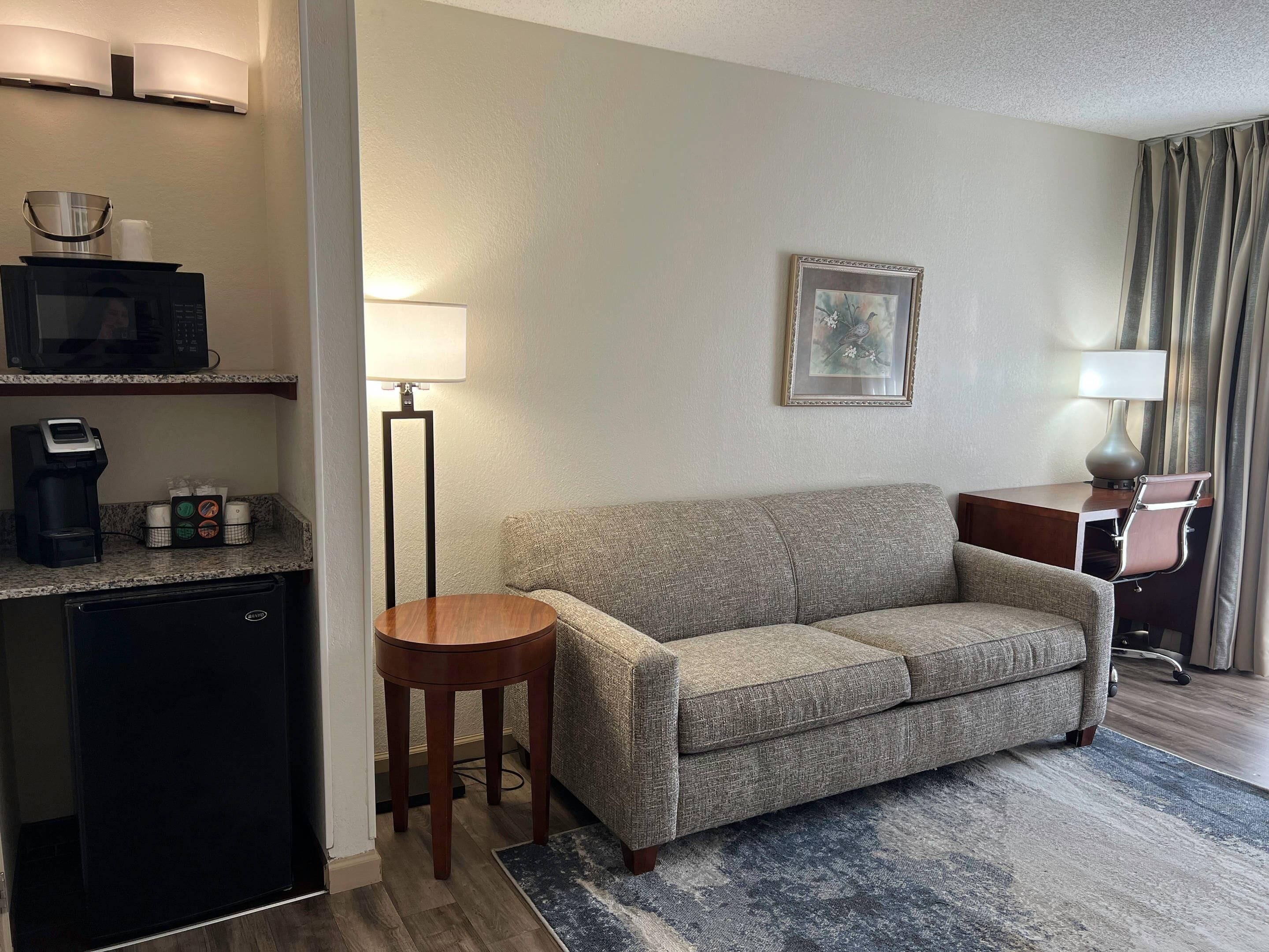 A hotel room with a sofa, a small table, a coffee machine, a lamp, and a picture on the wall.