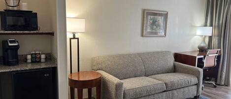 A hotel room with a sofa, a small table, a coffee machine, a lamp, and a picture on the wall.