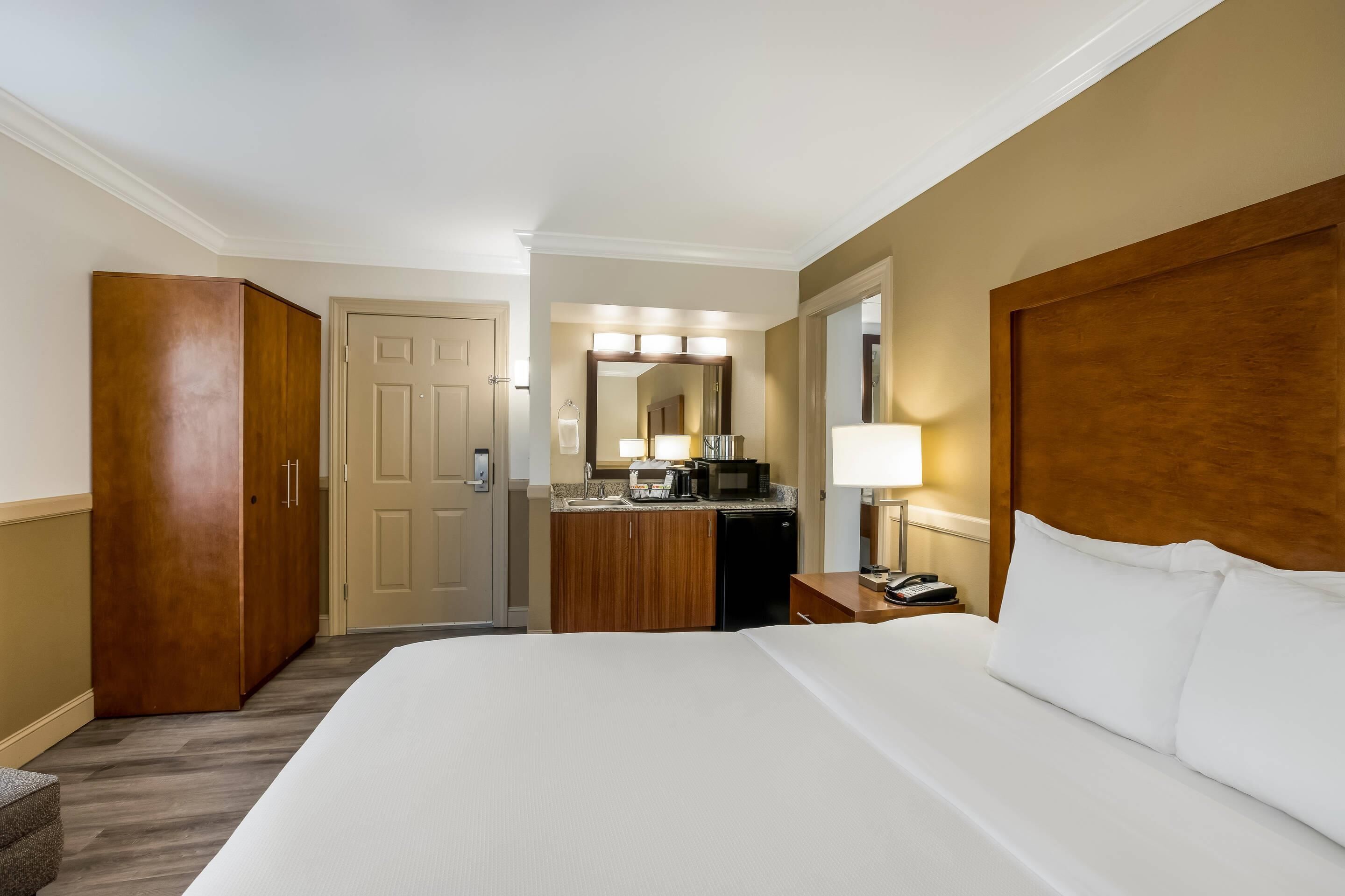 A hotel room with a large bed, a wooden wardrobe, a desk with a lamp, and a mirror above the mirror.