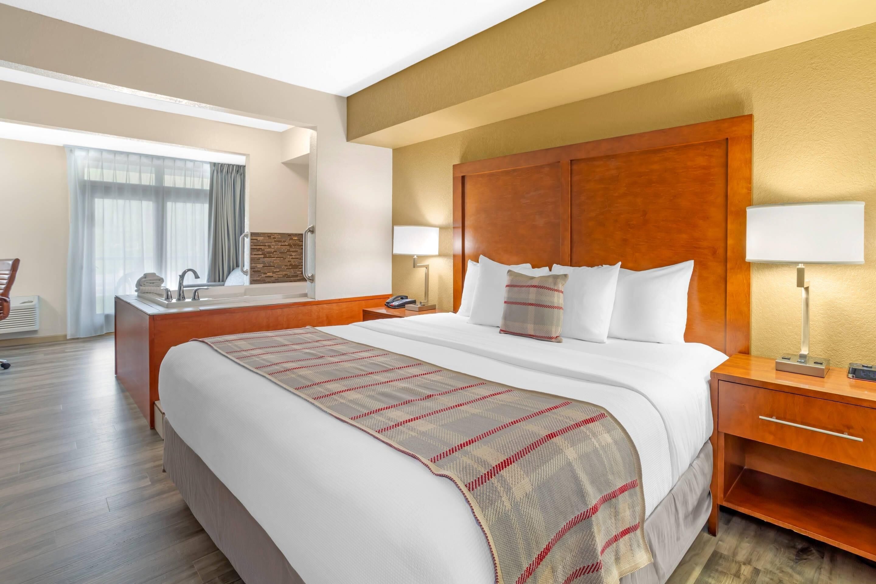 Hotelkamer met een groot bed, een houten hoofdeinde, een nachtkastje met lamp, een bureau met stoel, een raam met gordijnen en een spiegel aan de muur.