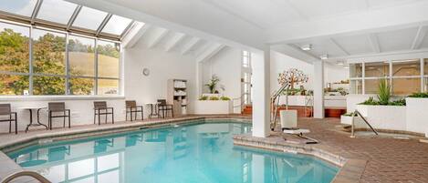 An indoor swimming pool with a view of trees, a sunroof, and a seating area with tables and chairs.