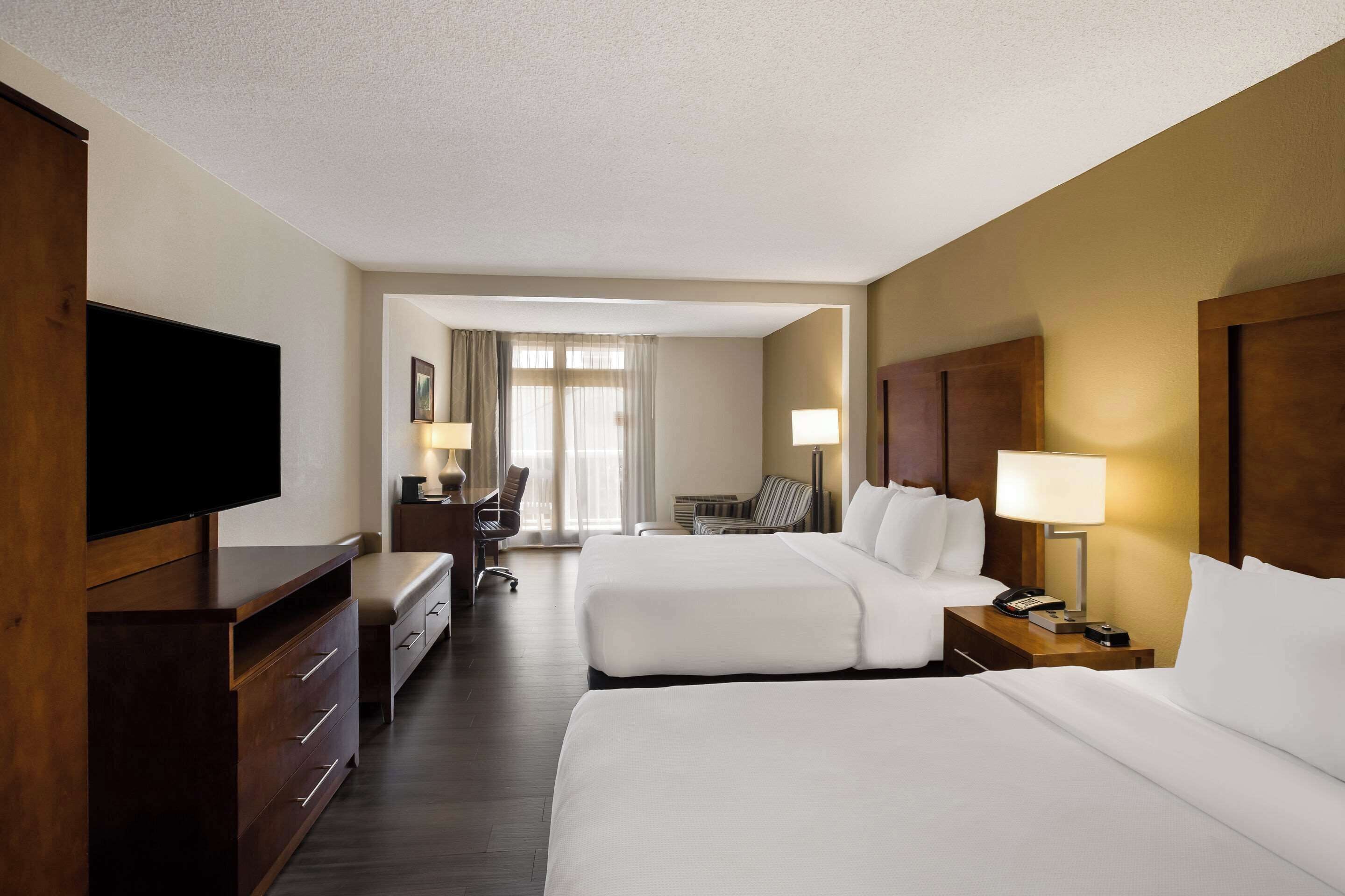 A hotel room with two beds, a desk, a chair, a television, and a lamp.
