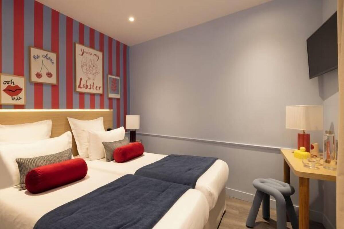 A hotel room with two beds, striped wallpaper, artwork on the wall, a desk with a lamp, and a TV mounted on the wall.