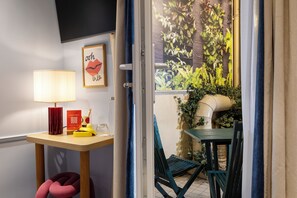 Une pièce comprenant un bureau, une chaise, une lampe, un tableau encadré et donnant sur un jardin avec des plantes et un tuyau.