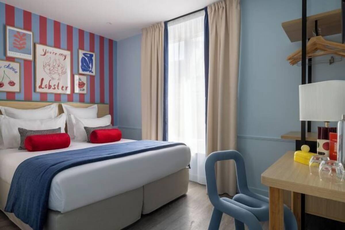 A hotel room with a bed, a chair, a table with a lamp, and a window with curtains.