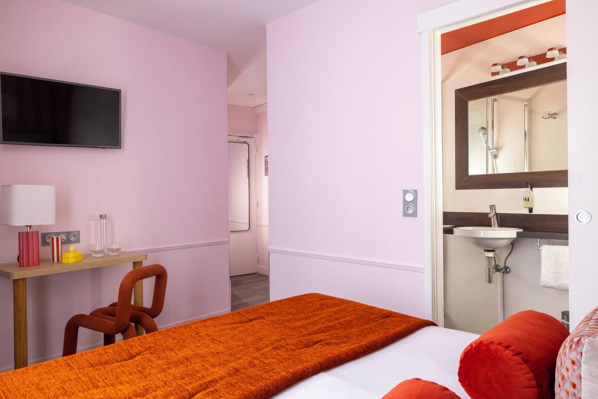 A hotel room with a bed, a desk with a chair, a TV, a bathroom with a sink and mirror.