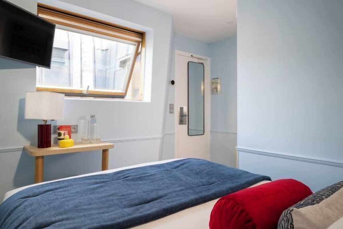 A bedroom with a bed, a bedside table with a lamp, a mirror, and a window with blinds.