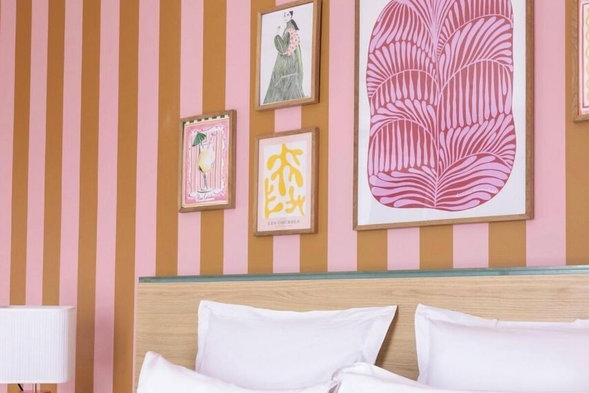 A hotel room with a striped wall, a bed with white and patterned pillows, and framed artwork on the wall.