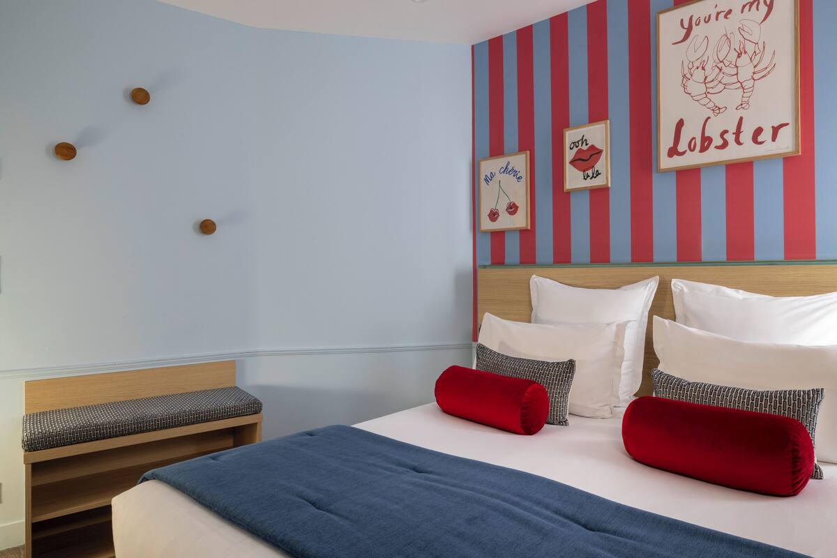 A neatly made bed with a blue blanket and red pillows, a wooden headboard, and striped wall decorations.