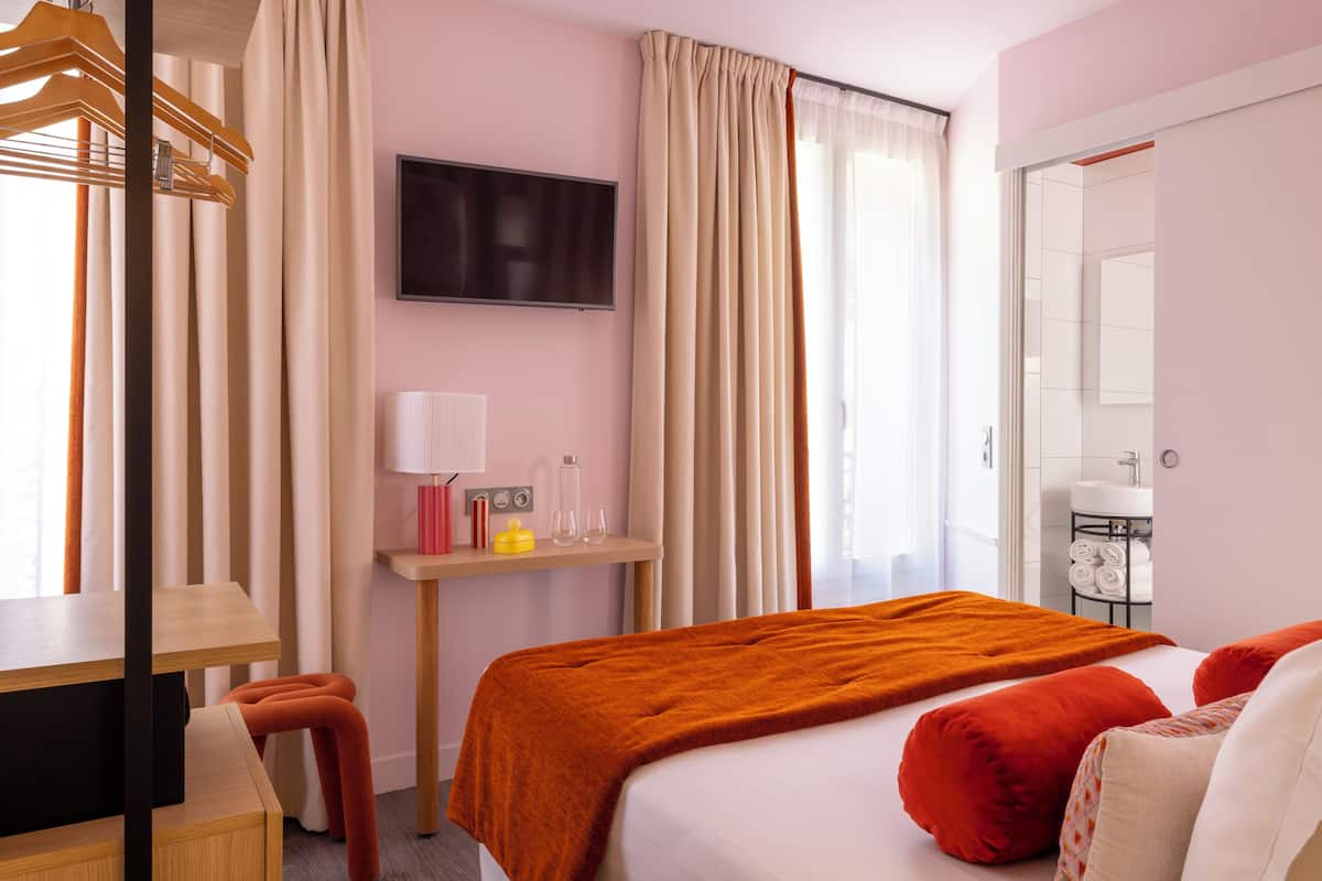 A hotel room with a bed, a desk, a chair, a TV, a window with curtains, and a bathroom visible through an open door.