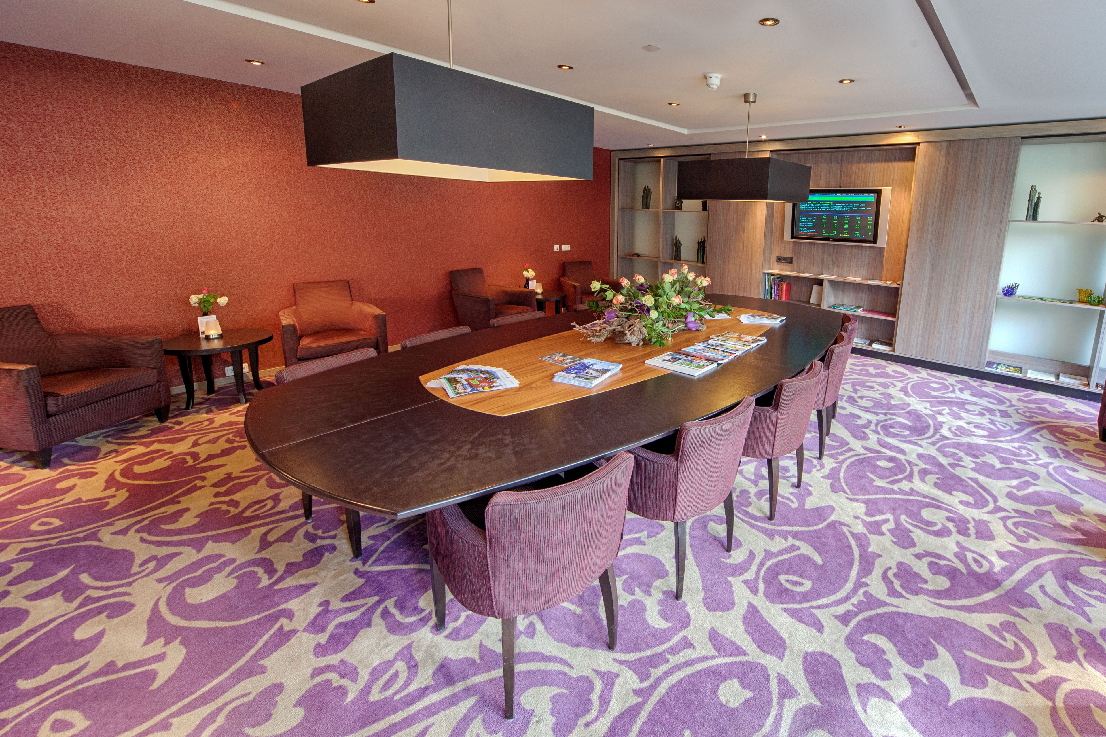 A conference room with a long table, six chairs, a TV screen, and a bookshelf.
