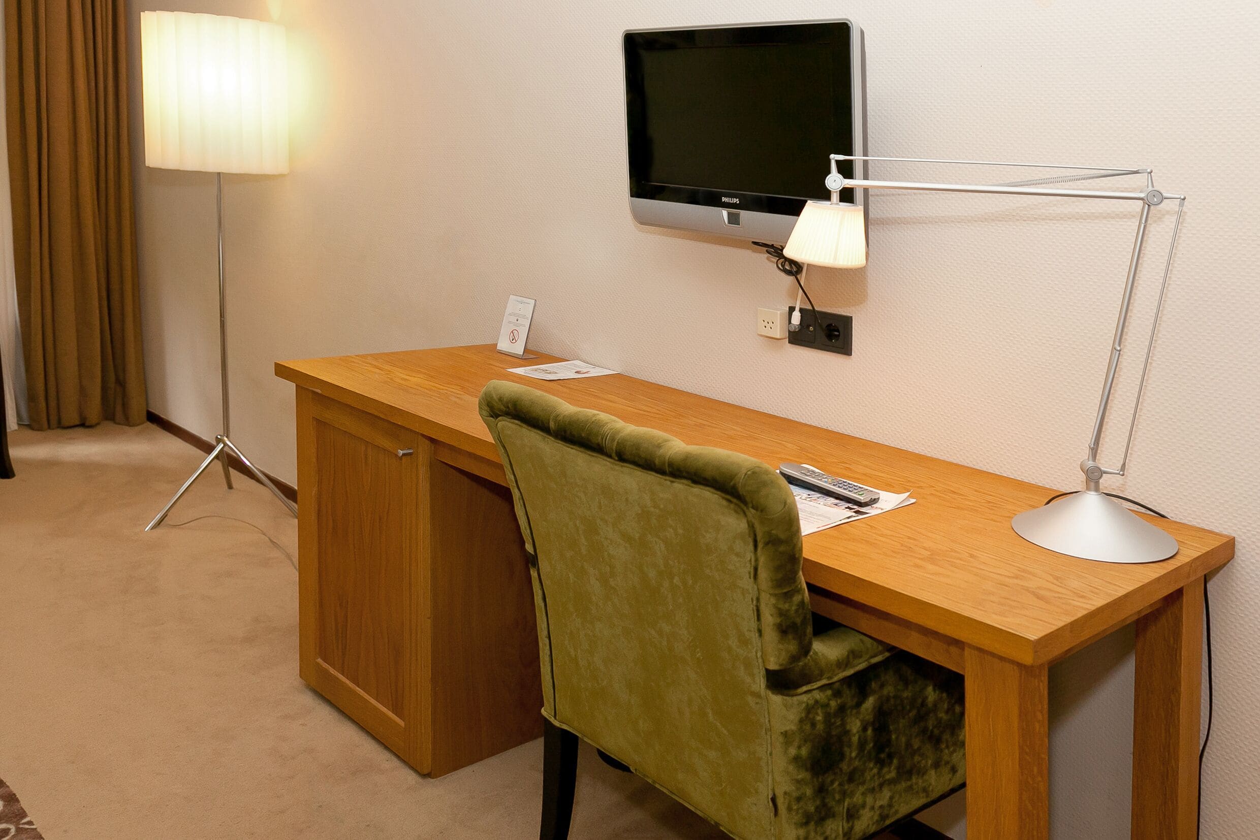 Hotelkamer met een houten bureau, een groene stoel, een flatscreen televisie aan de muur, een bureaulamp en een vloerlamp.