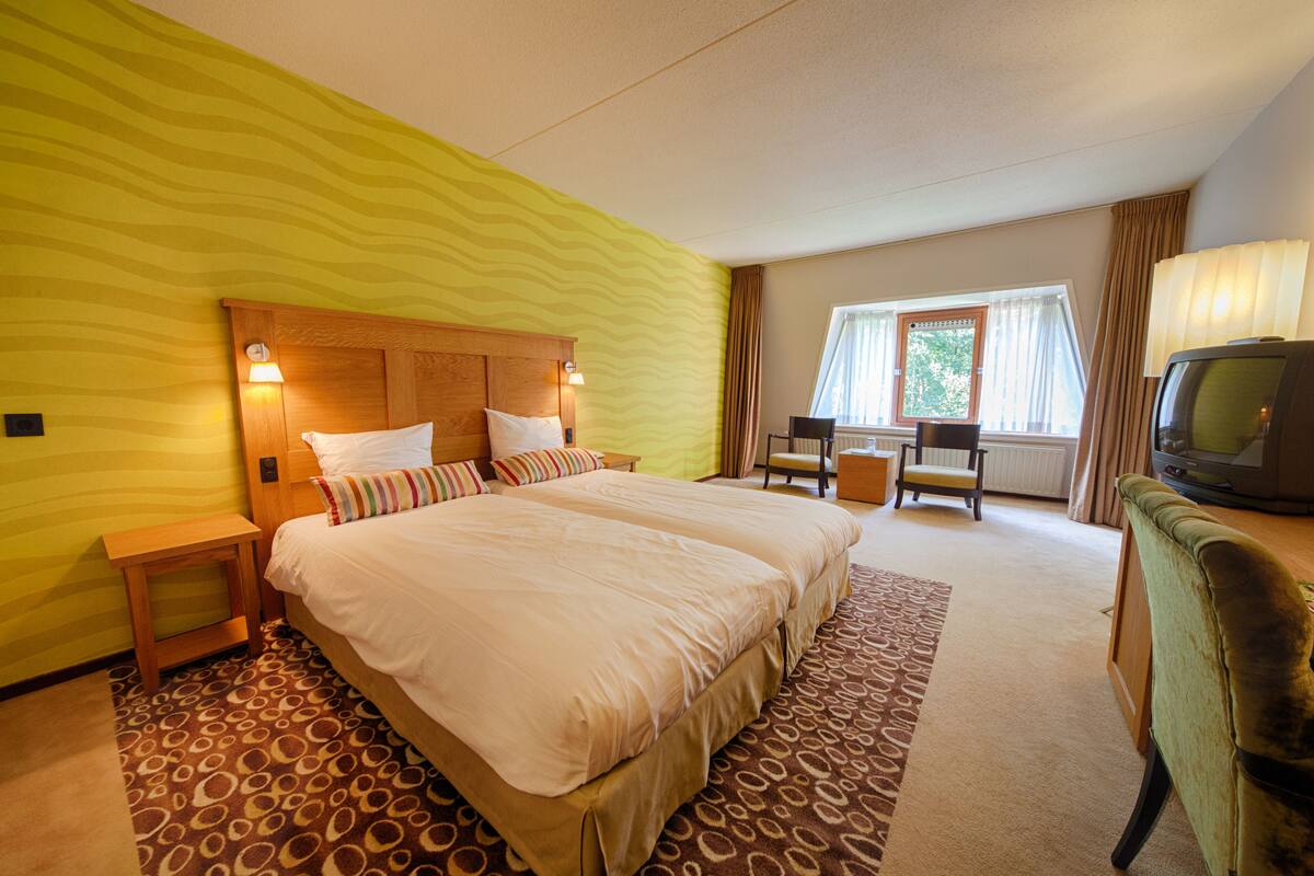Una habitación de hotel con una cama grande, un cabecero de madera, una televisión, un escritorio y una ventana con cortinas.