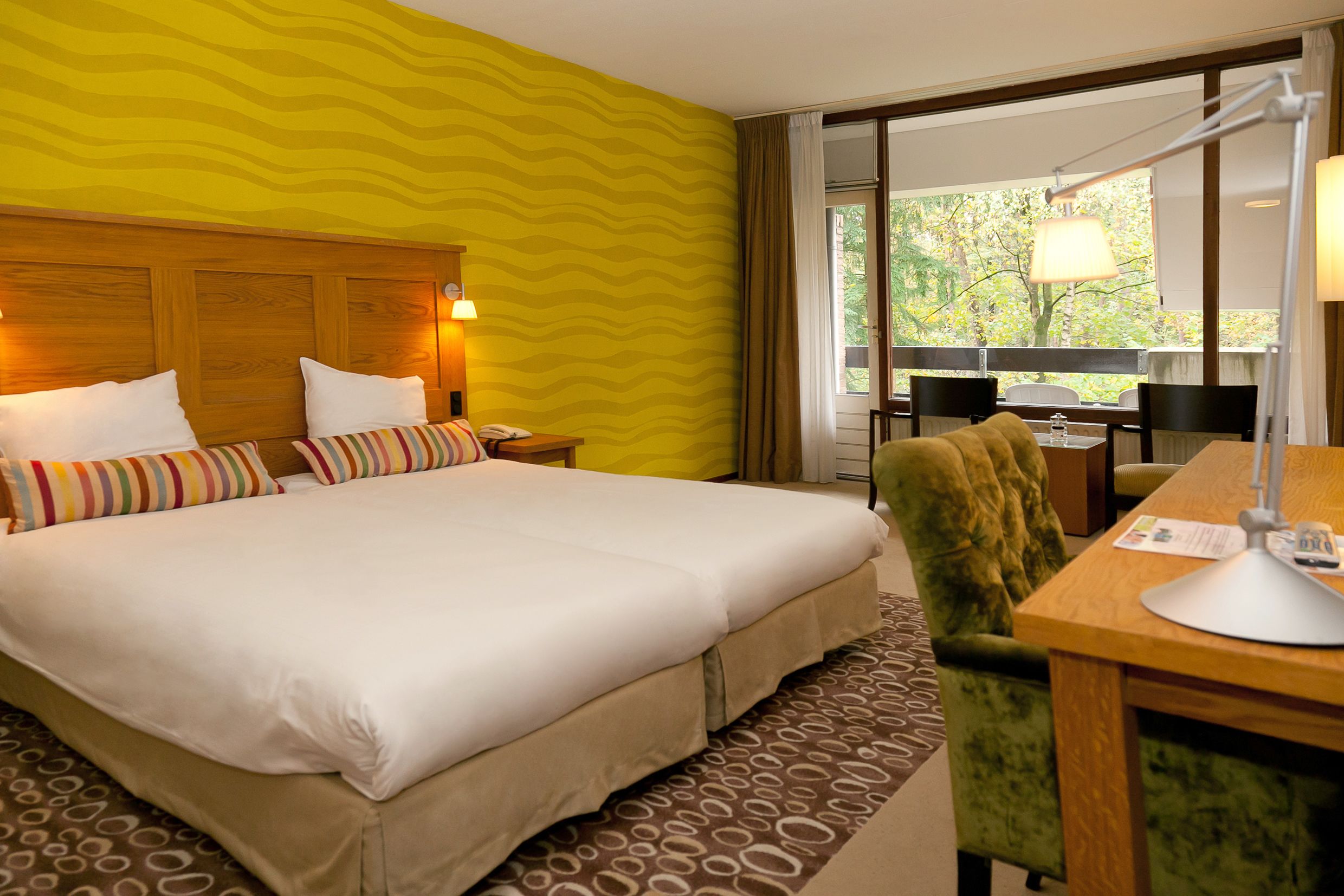 Habitación de hotel con una cama grande, un escritorio con lámpara, una silla y vistas a árboles por la ventana.