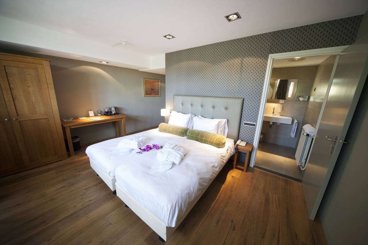 Un dormitorio ordenado con una cama grande, un armario de madera, una mesita de noche con una lámpara y un baño visible a través de una puerta entreabierta.