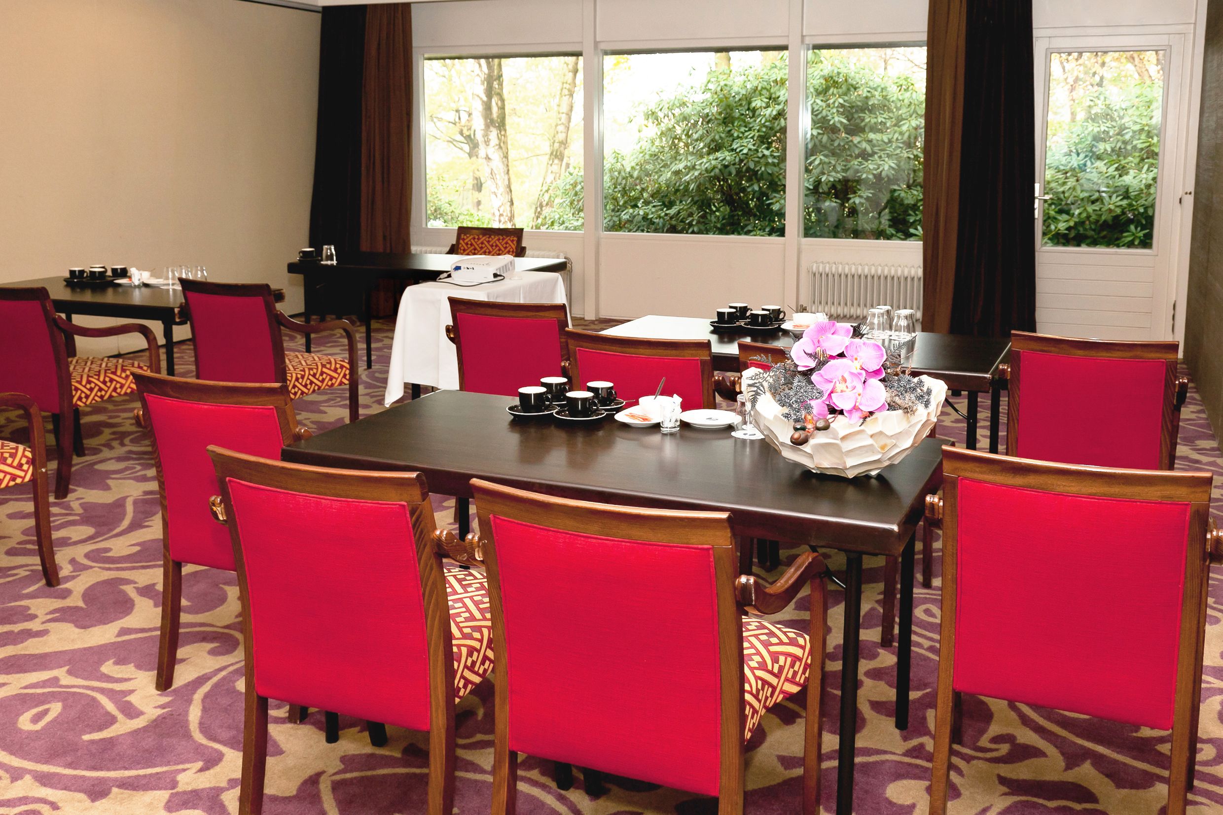 A conference room with a large table, red chairs, and a view of greenery through the windows.