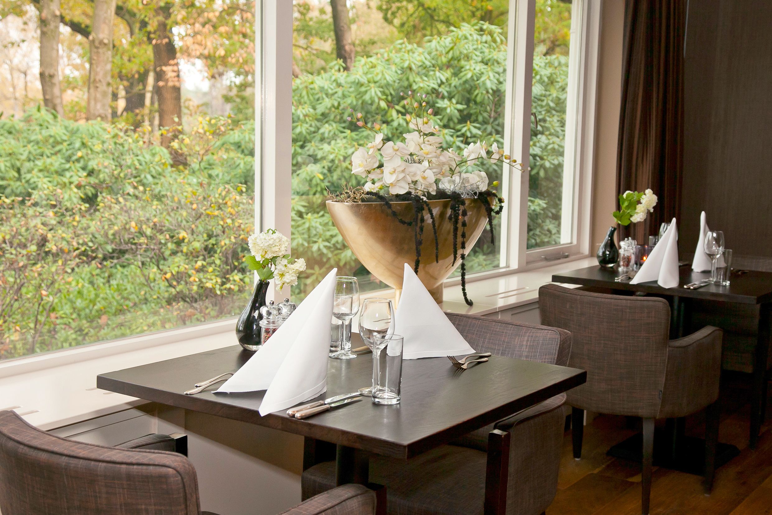 Un comedor con mesas dispuestas para dos, con servilletas blancas, cubiertos y cristalería. Un ventanal grande ofrece vistas a un jardín exuberante.