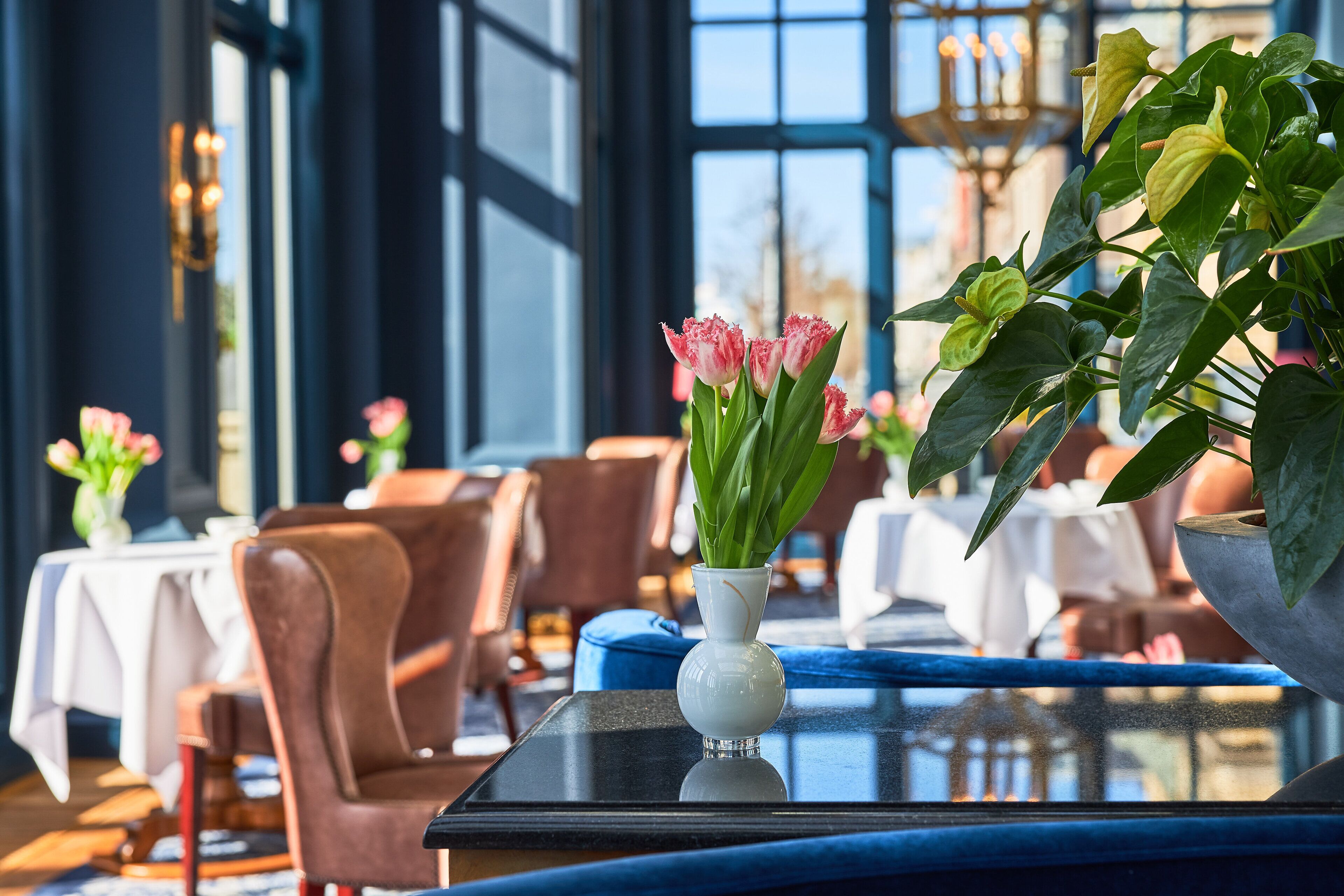 A dining area with tables and chairs, a vase with flowers, and a view through large windows.