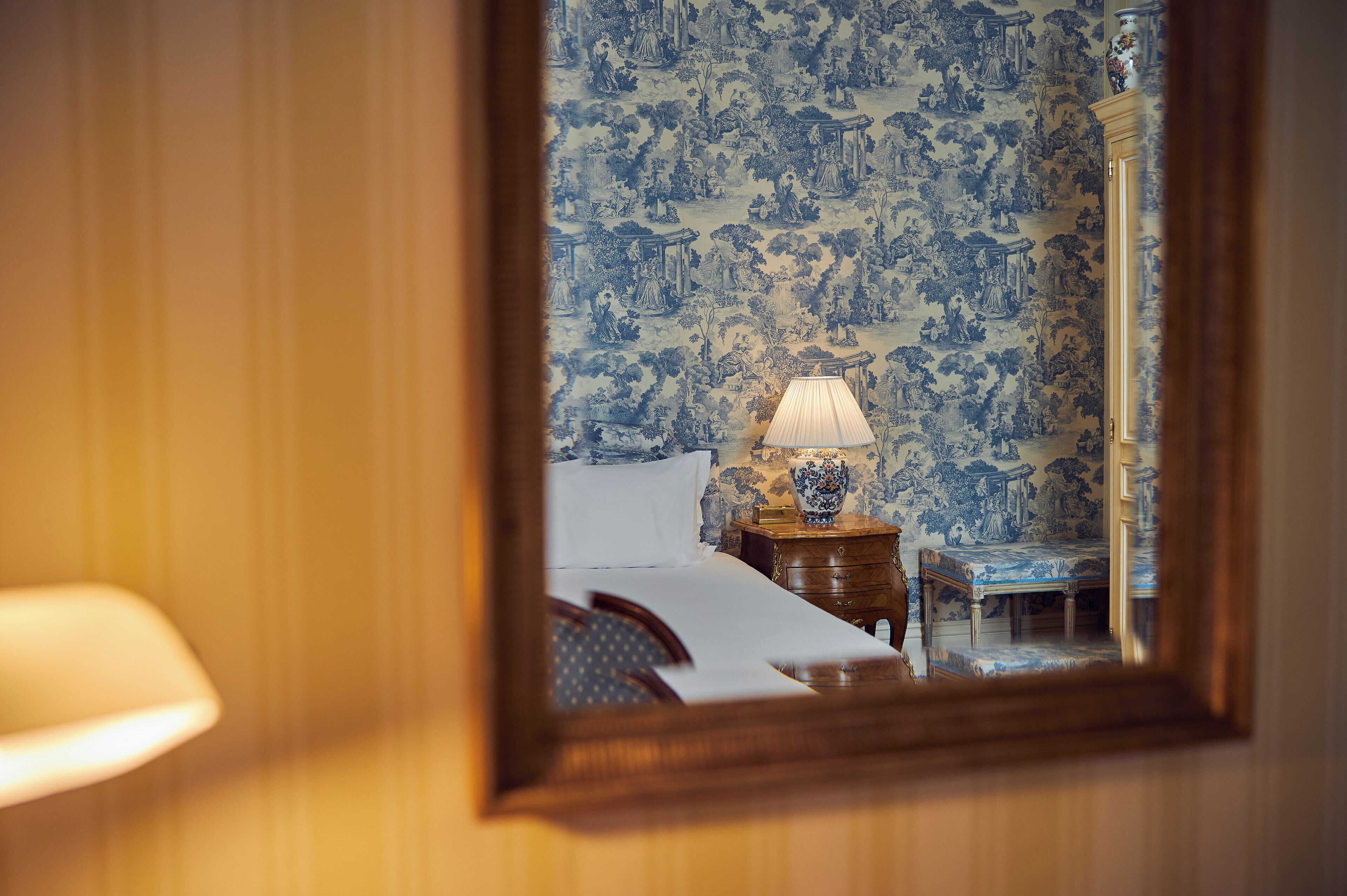 A room with a bed, a nightstand with a lamp, and a mirror reflecting a blue and white patterned wallpaper.