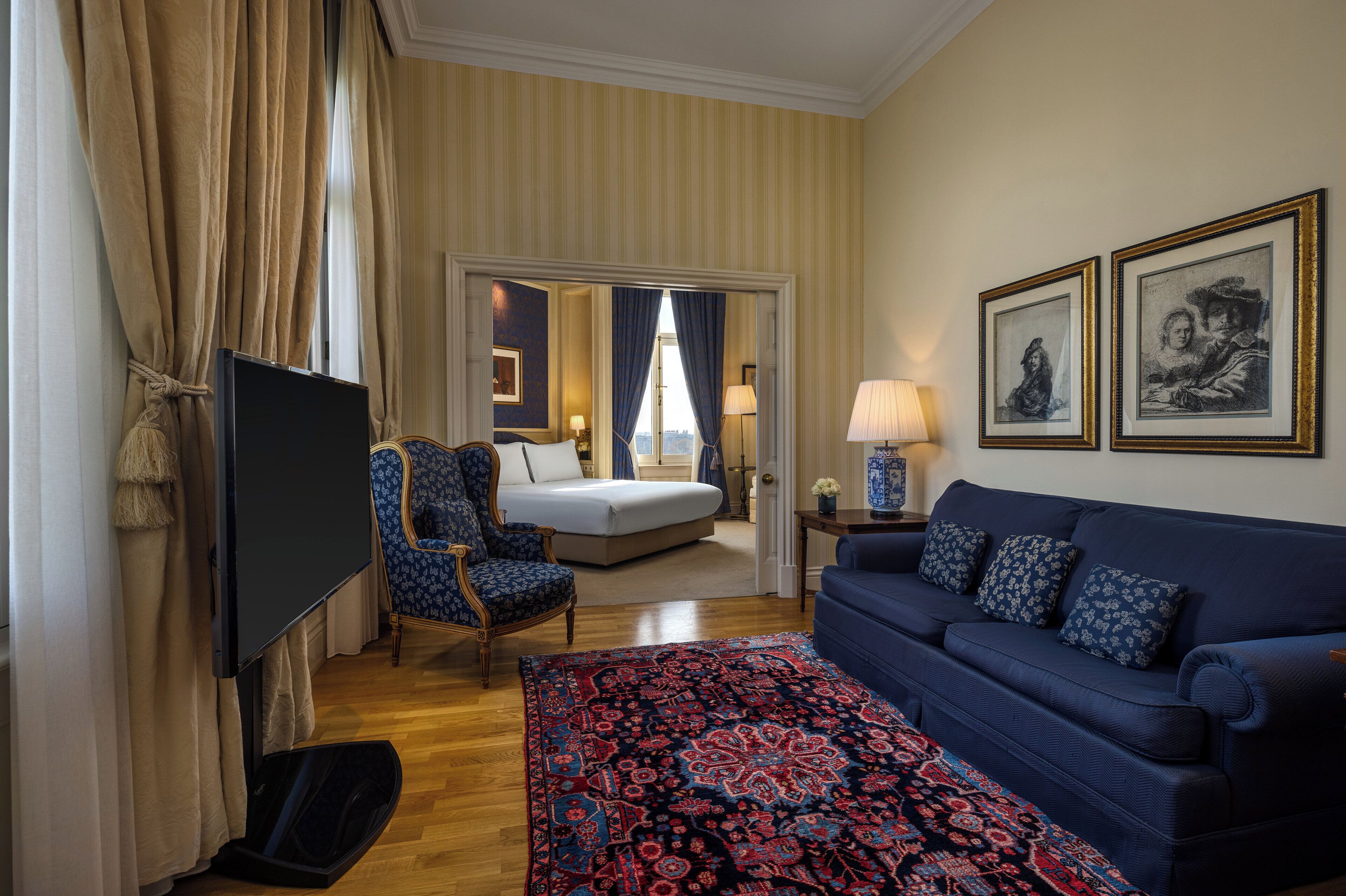 Una habitación de hotel con una cama, un sofá azul, un tapete estampado, un televisor y dos cuadros en la pared.