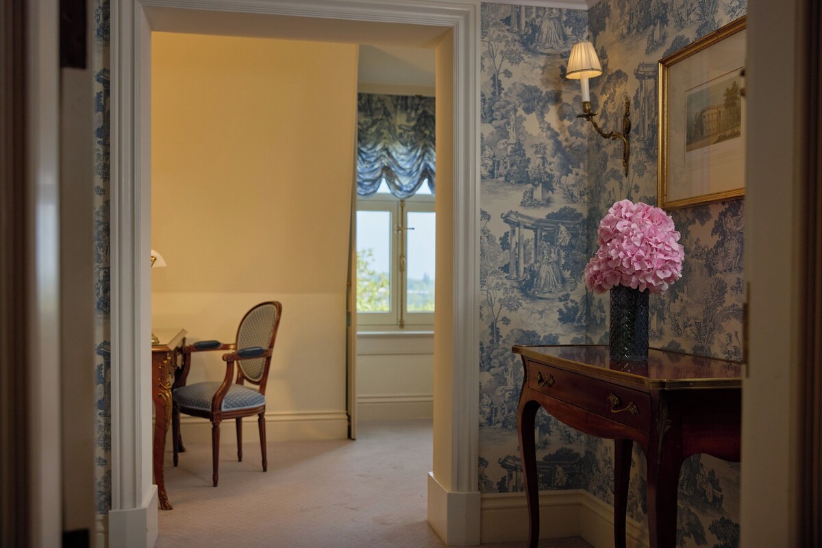 A room with a wooden chair, a desk with a vase of flowers, and a wall with a patterned wallpaper and a framed picture.