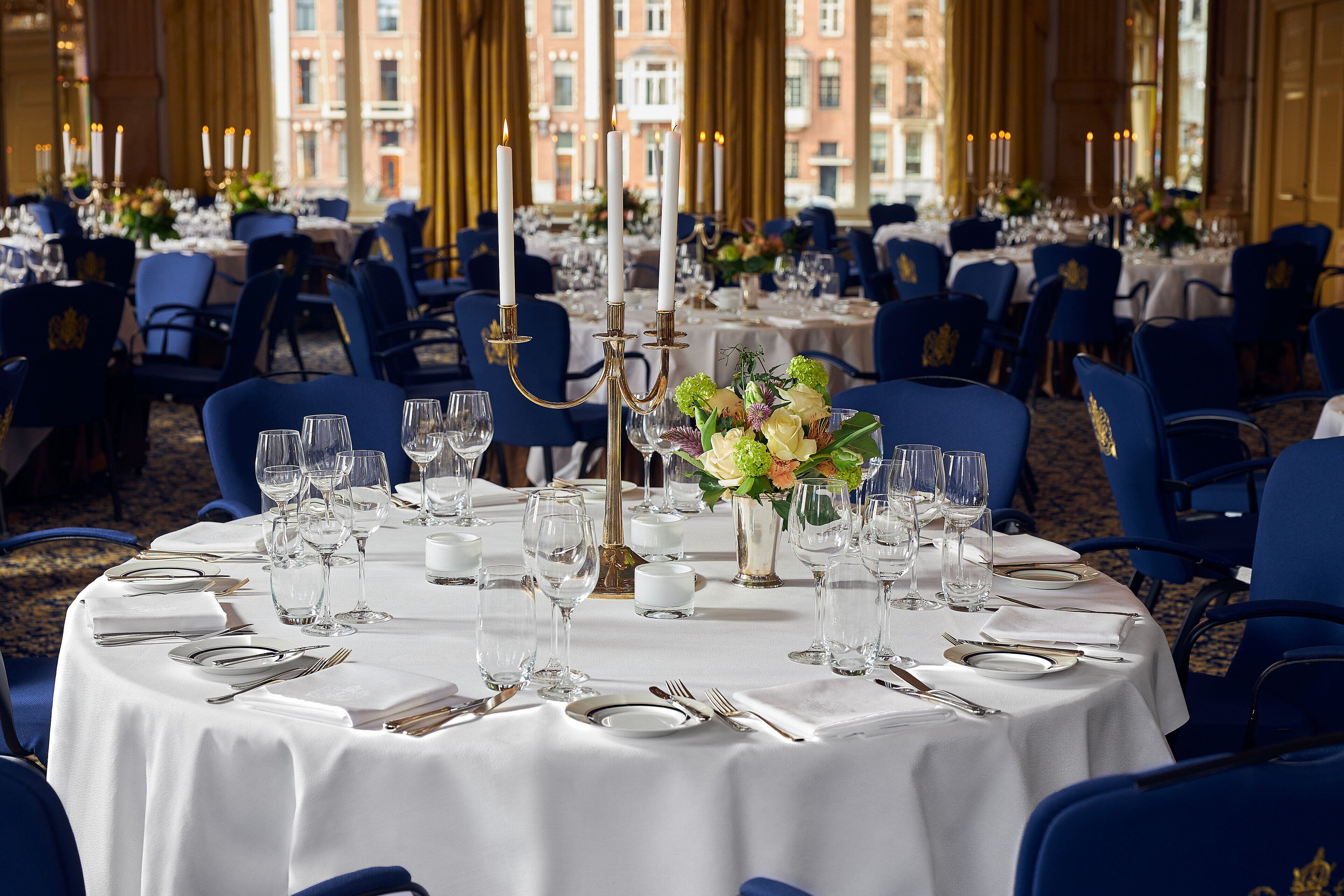 A formal dining room with round tables set for a meal, featuring white tablecloths, blue chairs, and candle centerpieces.
