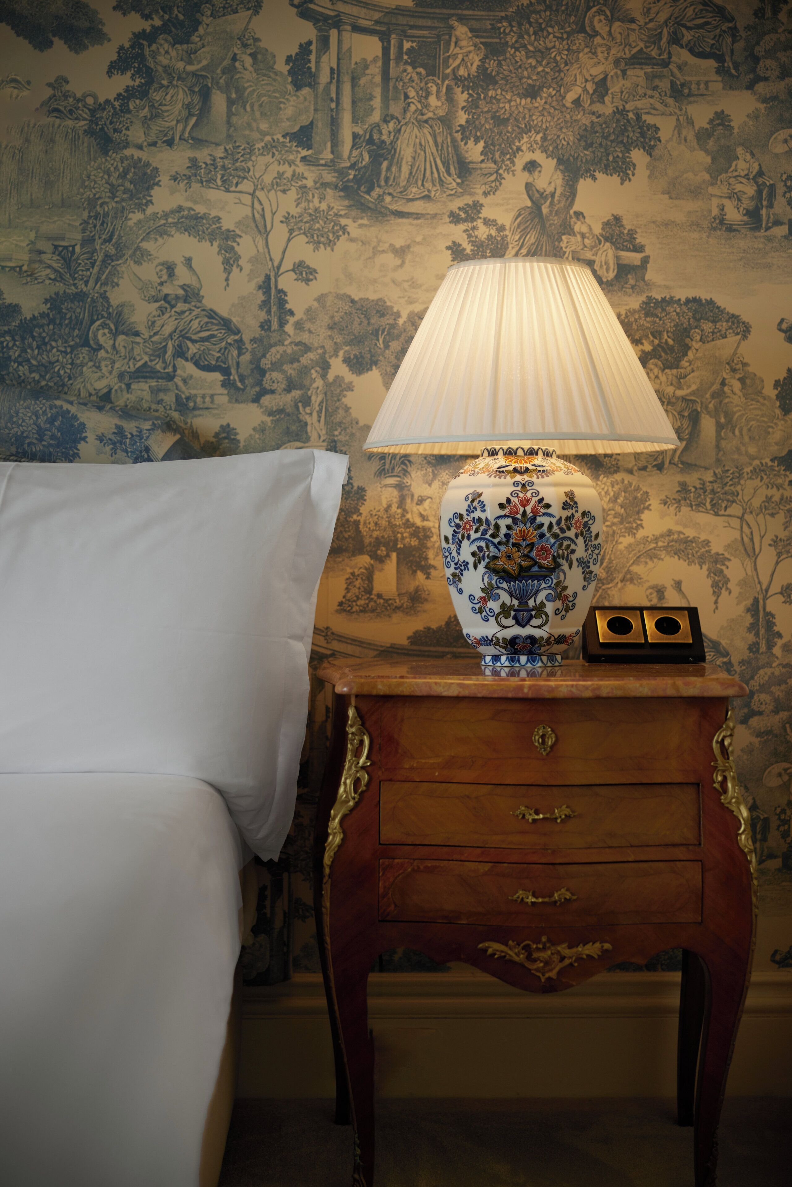 Een bed met witte lakens, een houten nachtkastje met een decoratieve lamp en een muur met een gedetailleerde muurschildering.