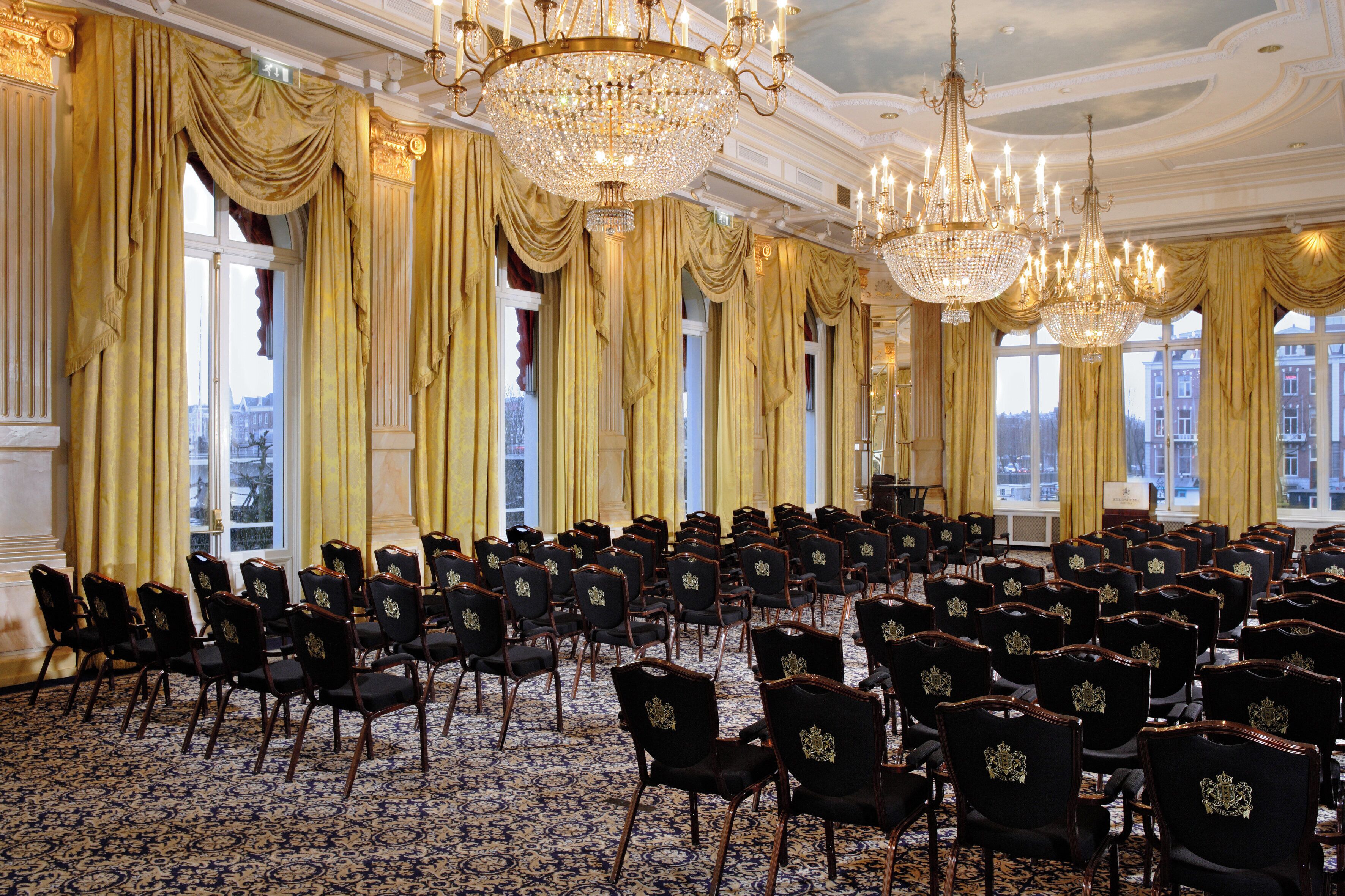 Un salón amplio con filas de sillas negras, candelabros decorados y grandes ventanales con cortinas.