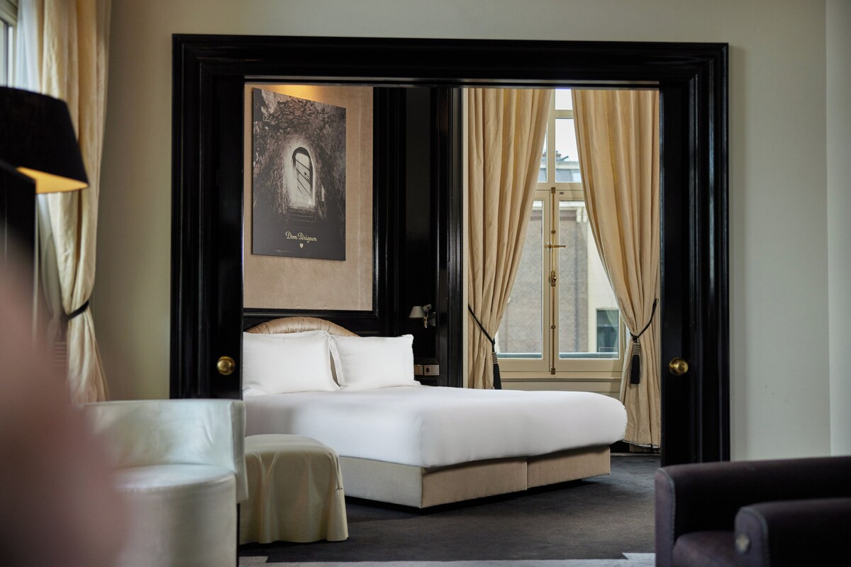 A hotel room with a bed, a window with curtains, a framed picture on the wall, and a chair.