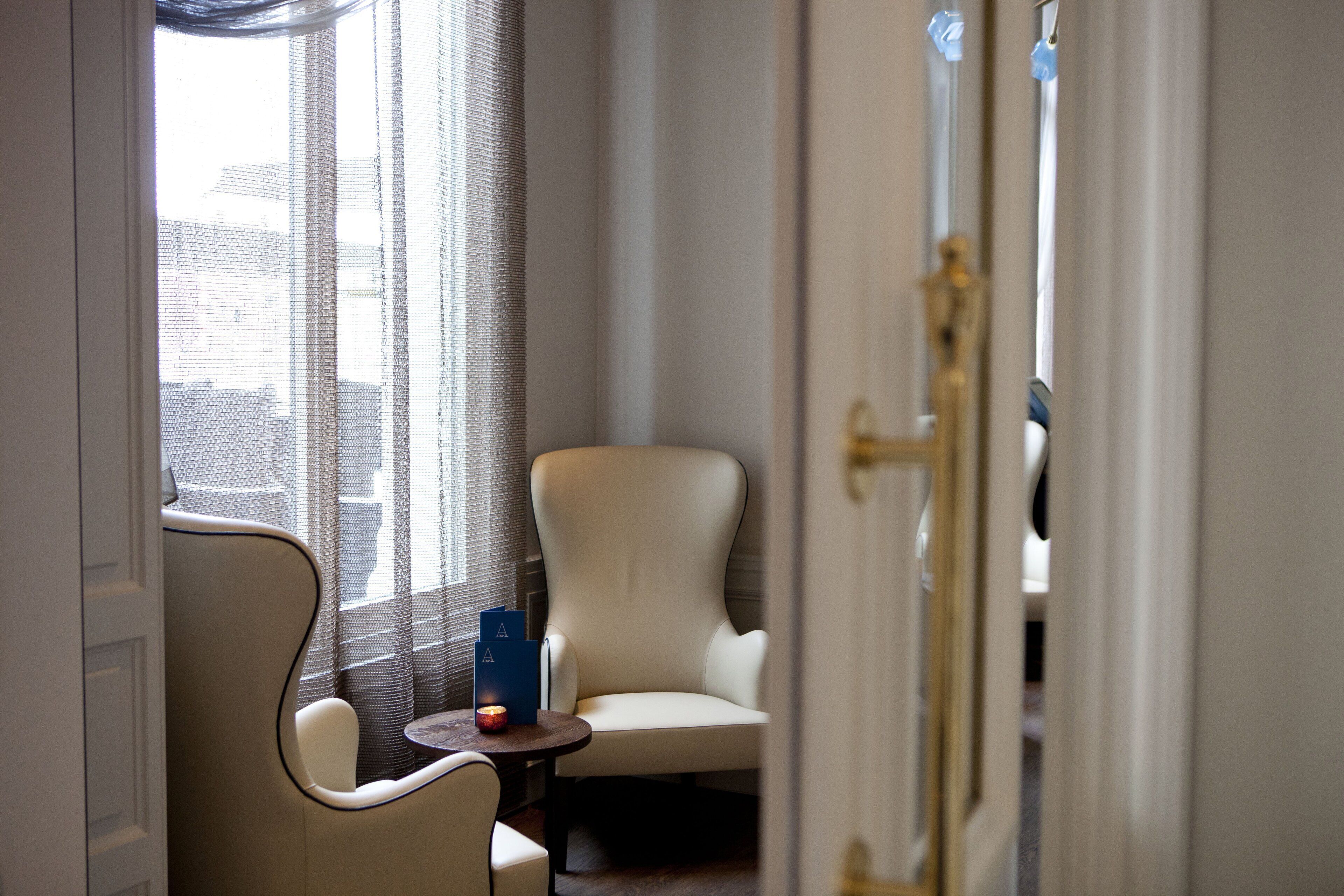 Ein Hotelzimmer mit einem großen Fenster, einem Stuhl, einem kleinen Tisch mit Buch und Kerze sowie einer Tür mit goldener Klinke.