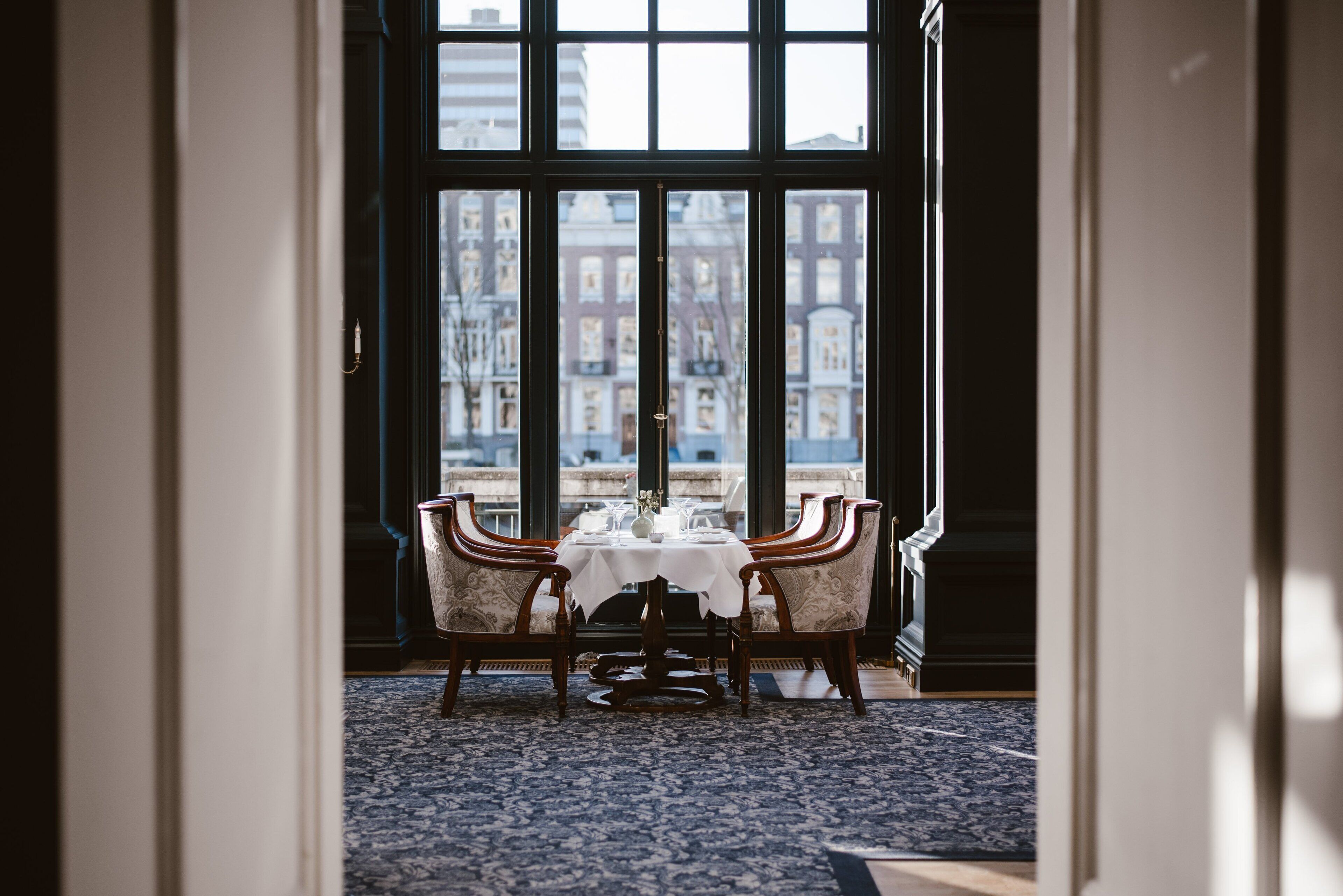 A dining area with a table set for two, large windows, and a view of buildings outside.