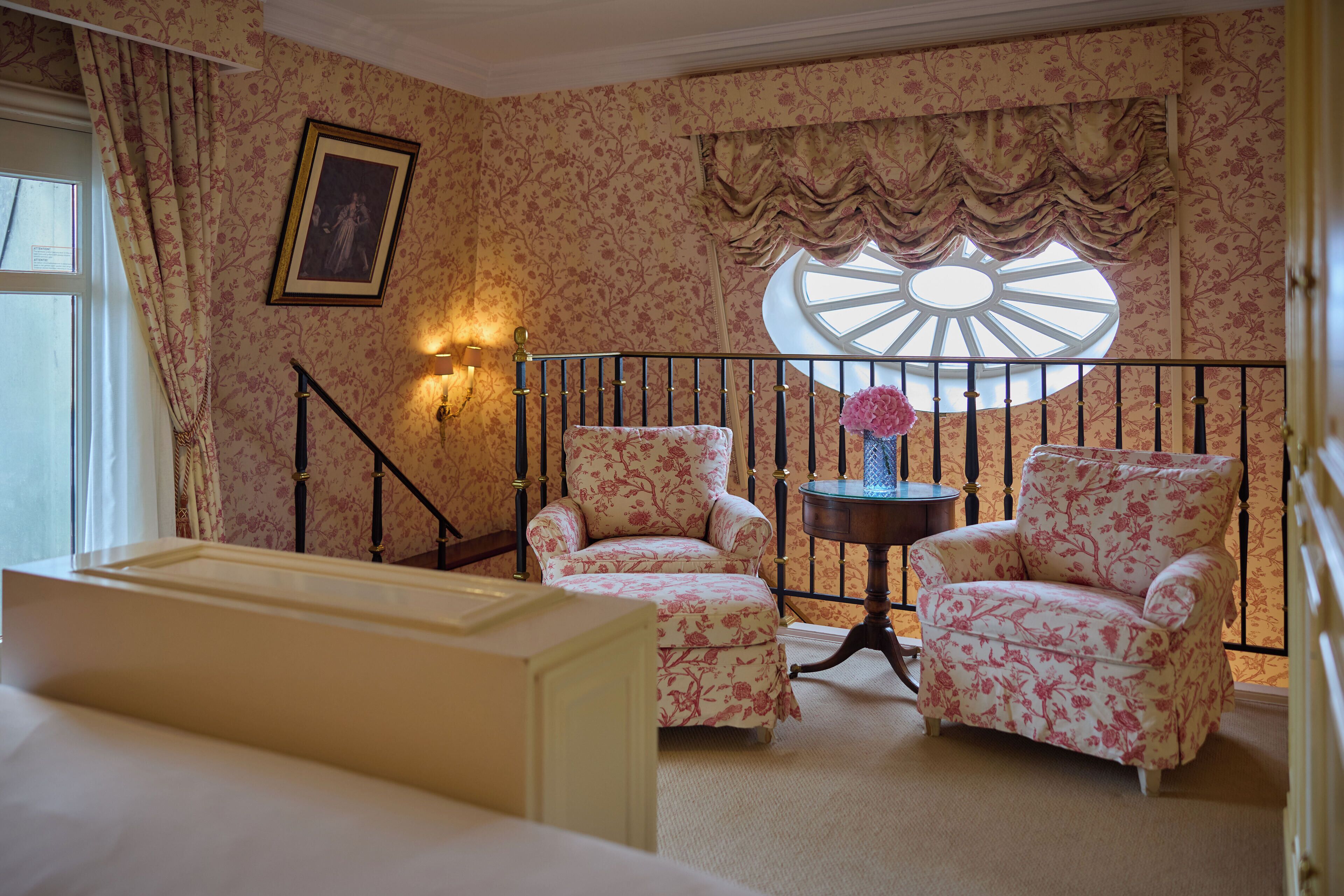 A room with floral wallpaper, two armchairs, a small table with a vase, a window with a decorative valance, and a staircase with a railing.