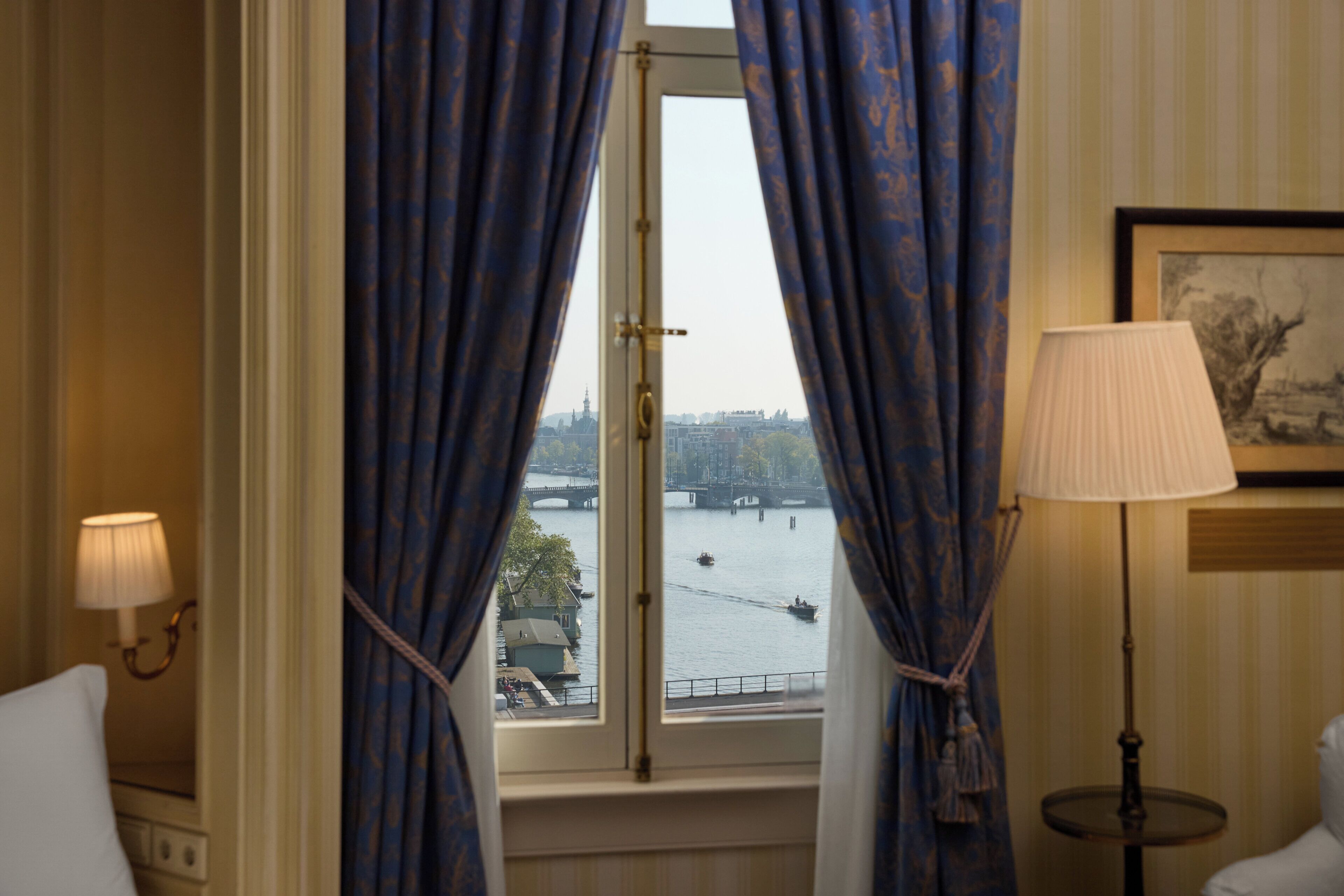 A room with a window overlooking a river, curtains, a lamp, and a framed picture on the wall.