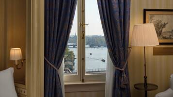 A room with a window overlooking a river, curtains, a lamp, and a framed picture on the wall.