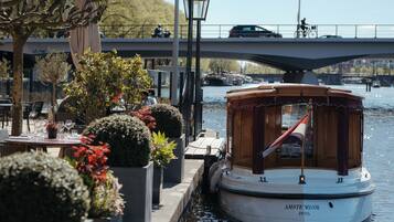 A boat docked at a riverside café with tables and chairs, a lamp post, and a bridge in the background.