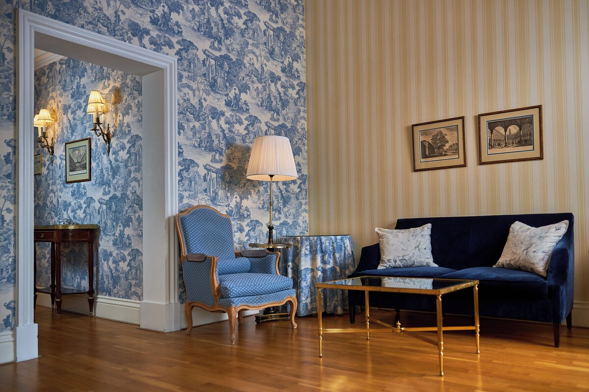 A room with a blue patterned wallpaper, a blue sofa with white pillows, a wooden chair, a small table, and two wall-mounted lamps.