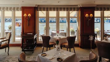 Un coin repas avec une table dressée pour deux personnes, de grandes fenêtres offrant une vue sur les bateaux sur l’eau et un meuble en bois avec une lampe.