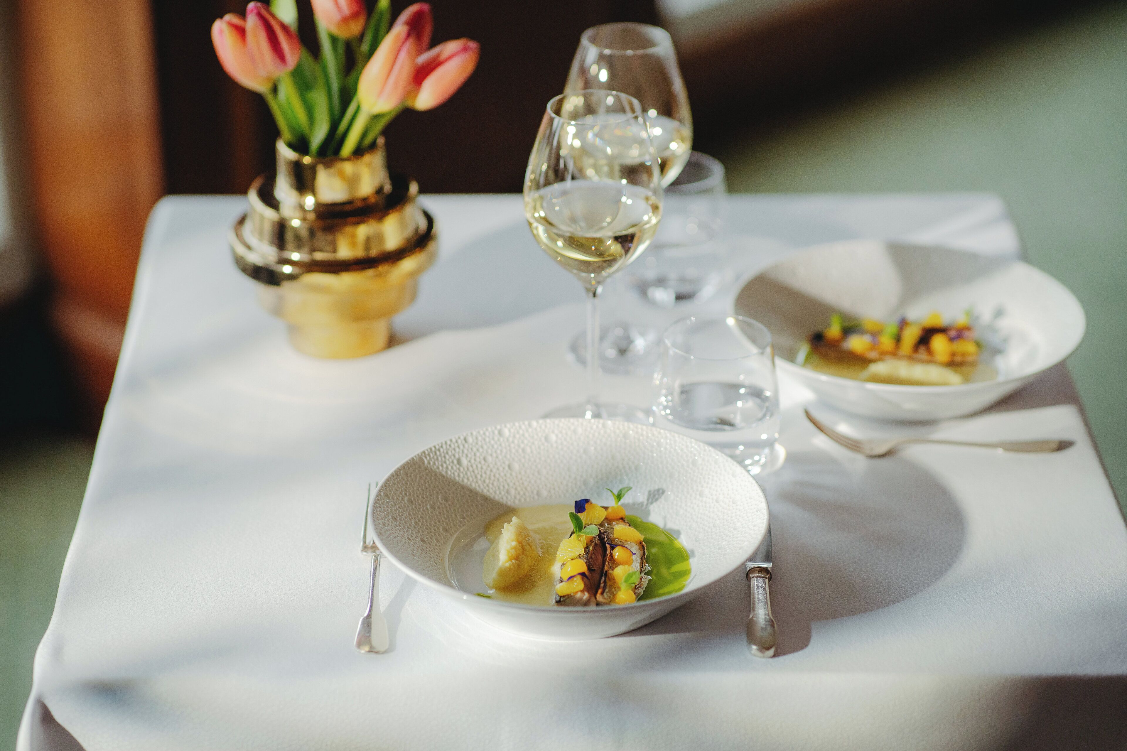 A dining table with a white tablecloth, a vase with pink tulips, and two glasses of white wine.
