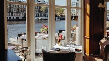 A dining area with a view of a river and buildings. There are tables set with wine glasses, plates, and cutlery.