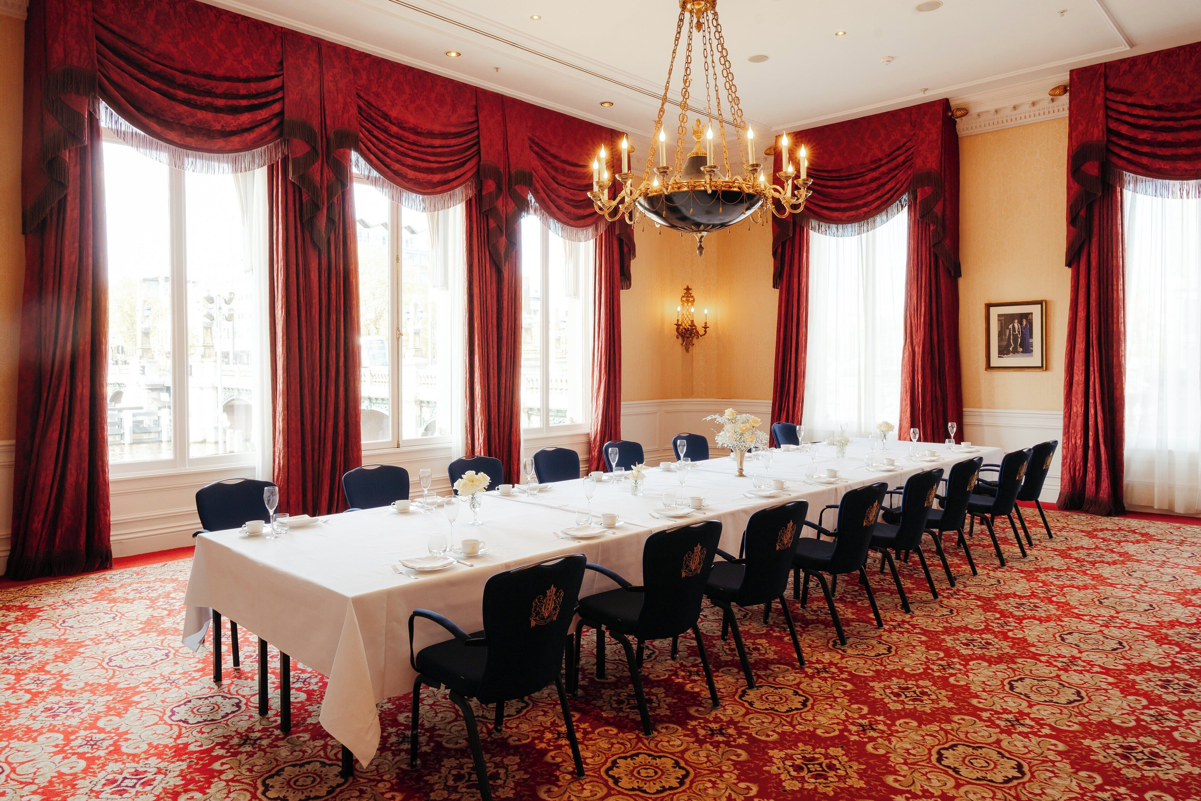 Un comedor formal con una larga mesa dispuesta para un banquete, cortinas rojas y una araña.