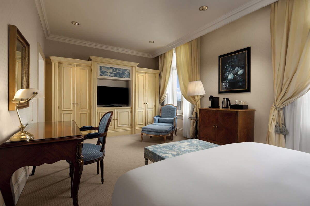 A hotel room with a bed, a desk with a lamp, a chair, a television, and a cabinet.