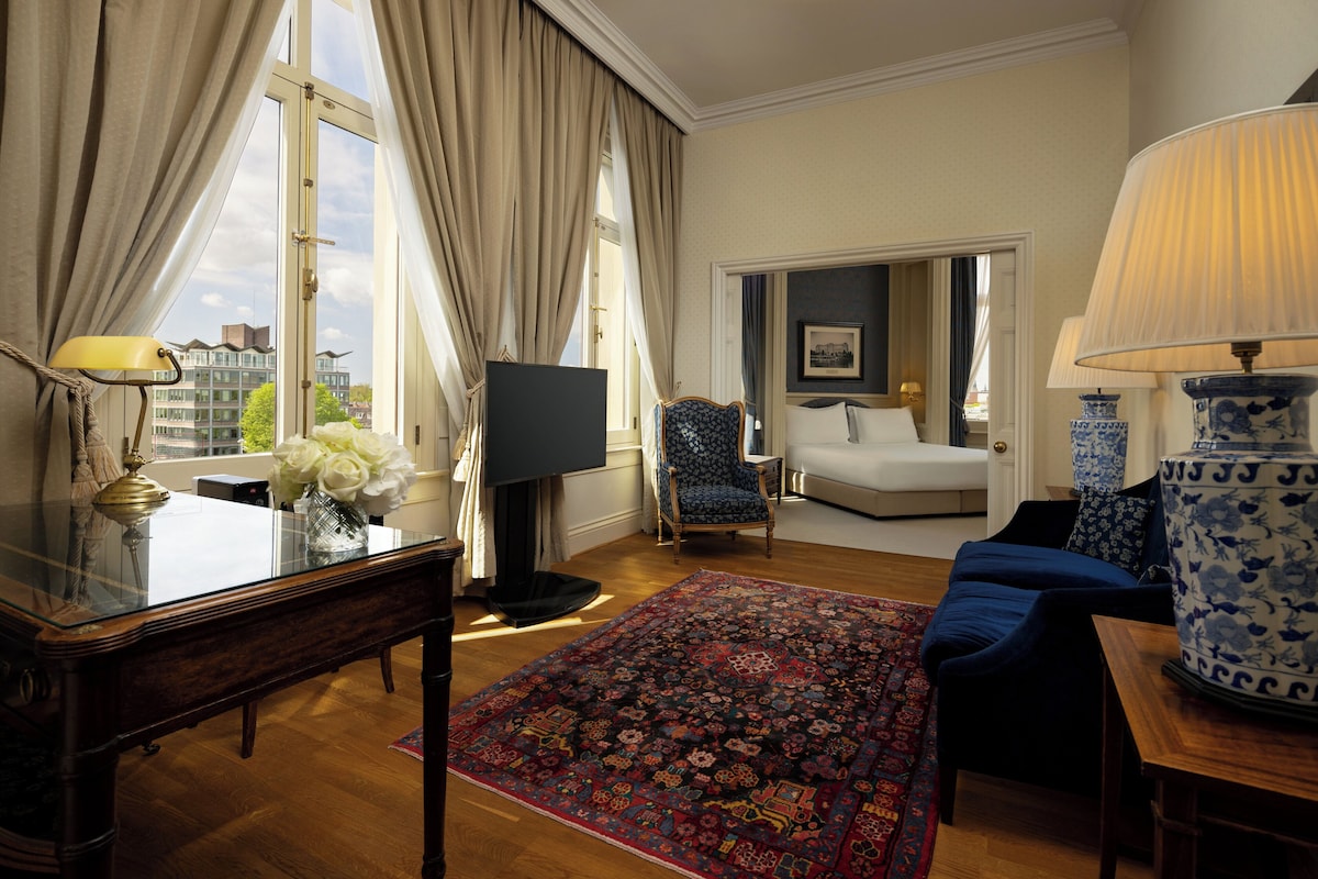 A hotel room with a large window, a bed, a blue sofa, a wooden table, and a decorative vase.