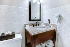 A bathroom with a white toilet, a sink with a mirror above it, and a hairdryer mounted on the wall. - Clarion Hotel & Suites (Metairie)