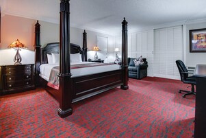 Una cama con dosel de madera oscura, dos mesitas de noche con lámparas, un escritorio con silla y una alfombra estampada.