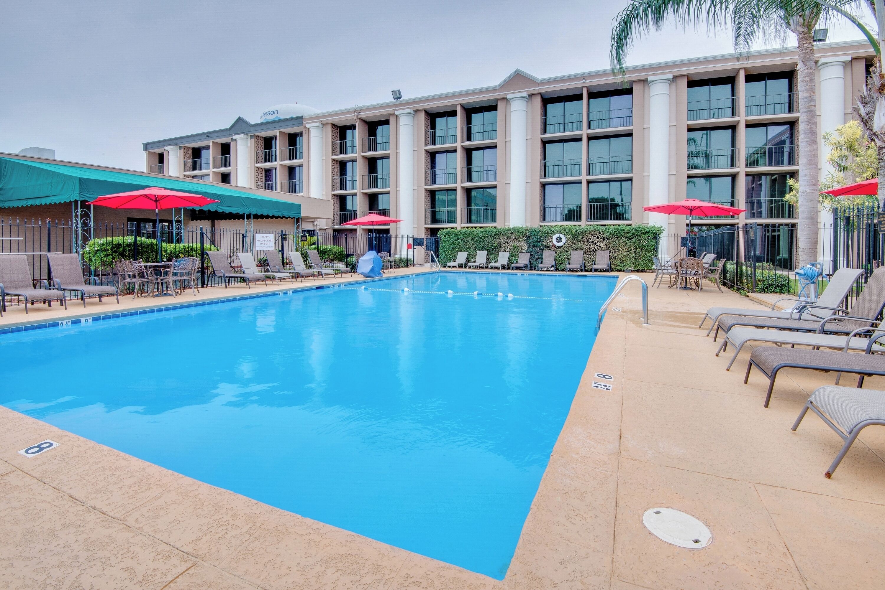 A swimming pool with lounge chairs and umbrellas, surrounded by a building with multiple floors and large windows.