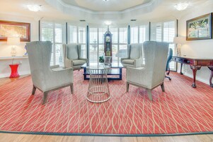Una habitación con alfombra roja estampada, una mesa redonda con sillas y ventanas grandes con persianas.