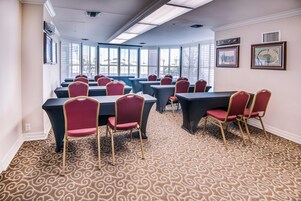 Un salón de conferencias con mesas y sillas, ventanas amplias, alfombra estampada y cuadros en las paredes.