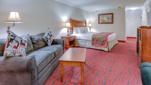 A hotel room with a grey sofa, a wooden coffee table, a bed with a red and green patterned bedspread, and a wooden nightstand with lamps. - Clarion Hotel & Suites (Metairie)